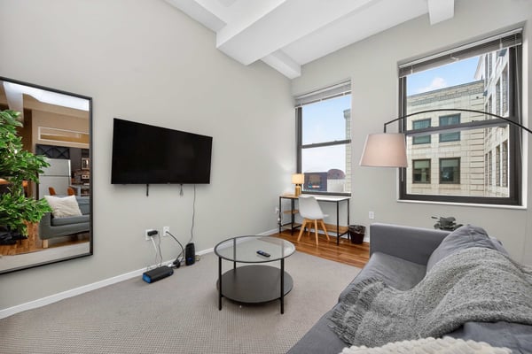 Modern living room with gray sofa, wall-mounted TV, round glass coffee table, and large windows overlooking city buildings