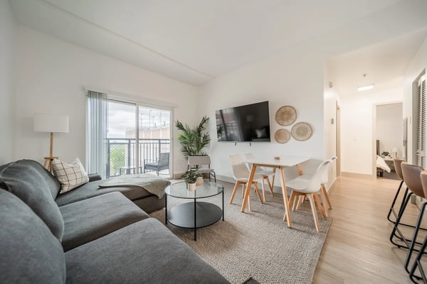 Modern open-plan living room with gray sectional sofa, dining table, wall-mounted TV, and balcony overlooking city skyline.
